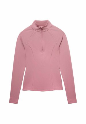 Kvinde iført mauve træningstøj med en top med halv lynlås, matchende shorts, løs mauve jakke, sort jakke bundet omkring taljen og sorte sneakers.