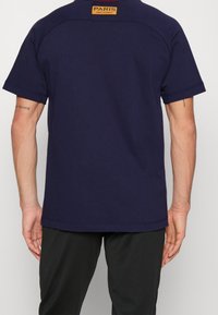 Camiseta de manga corta azul marino con un escote redondeado, que presenta una etiqueta rectangular naranja en la parte posterior con "PARIS SAINT-GERMAIN."