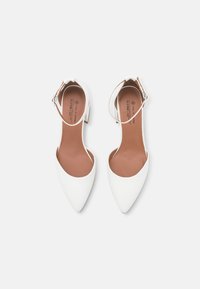 Escarpins blancs à bout pointu avec une fine bride de cheville. Surface lisse et semelle intérieure beige avec logo. Petit talon empilé pour une légère élévation.