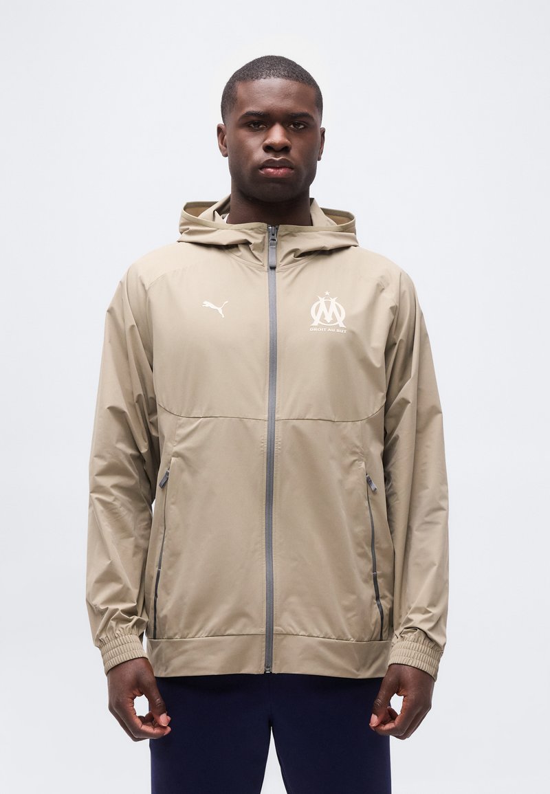 Άνδρας που φοράει μπεζ φούτερ με φερμουάρ και κουκούλα, με λογότυπα Puma και Olympique de Marseille, στέκεται μπροστά σε απλό λευκό φόντο.