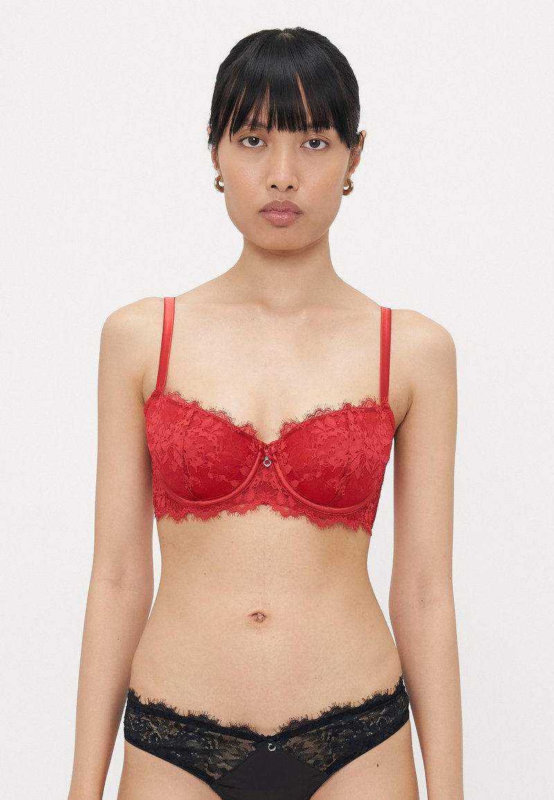 Red lace bra with underwire and thin adjustable straps. Features decorative scalloped edges and a central embellishment. Black lace briefs included.