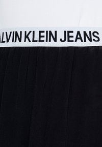 Tessuto plissettato nero con una vita bianca che presenta la scritta "CALVIN KLEIN JEANS" in lettere nere. Il tessuto appare liscio e morbido.