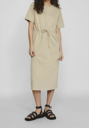 Robe beige longueur genou avec des manches courtes et une taille à cordon. Assortie à des sandales noires à brides avec des accents métalliques.