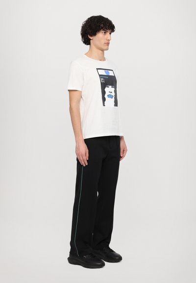 White cotton t-shirt featuring a graphic print with blue accents, paired with black pants that have a teal side stripe. Black sneakers.