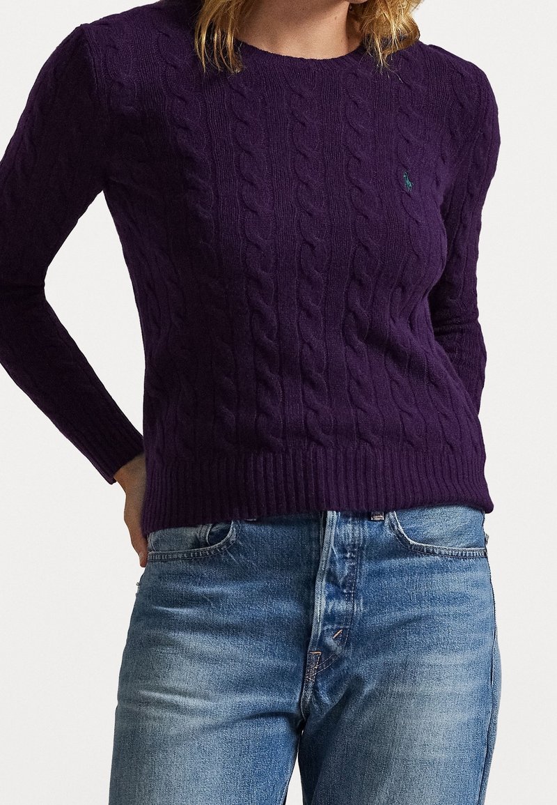 Femme portant un pull en tricot torsadé violet foncé et un jean bleu, debout avec une main sur la hanche devant un fond uni.