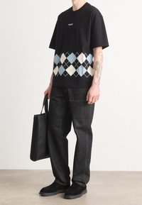 Schwarzes Baumwoll-T-Shirt mit einem blau-weißen Rauten-Muster am Saum, kombiniert mit dunklen Denim-Hosen und schwarzen Schuhen. Hält eine schwarze Tragetasche.