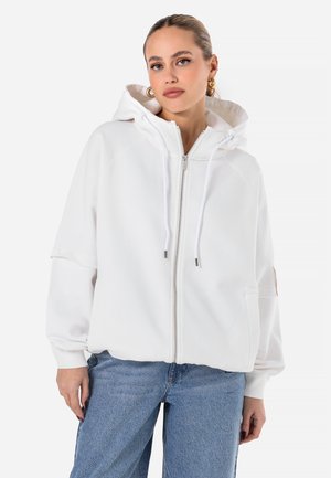 White zip-up hoodie with a drawstring hood, ribbed cuffs, and two front pockets. Worn with light blue denim jeans, featuring a relaxed fit.