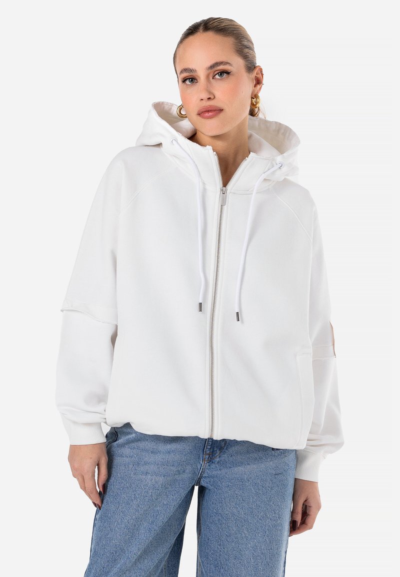 Felpa bianca con zip e cappuccio con cordino, polsini a coste e due tasche frontali. Indossata con jeans di denim blu chiaro, dal fit rilassato.