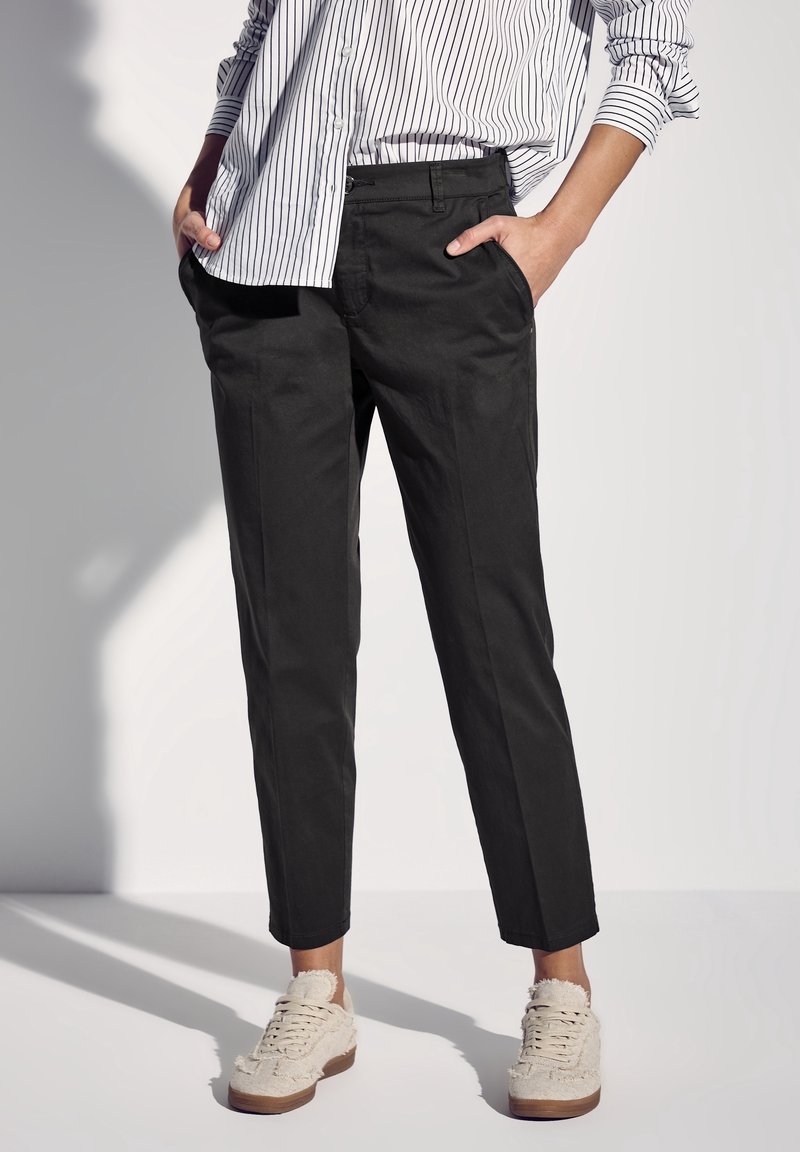 Personne portant un pantalon noir fuselé, des baskets blanches en tissu texturé, et une chemise blanche à manches longues avec des rayures verticales noires, les mains dans les poches.