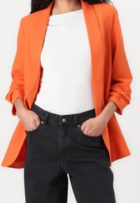Blazer naranja de corte relajado, con solapas de muesca y mangas enrolladas. Combinado con una camiseta blanca y jeans negros de talle alto.