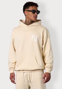 Sweatshirt beige à capuche avec une poche frontale, présentant un logo blanc sur la poitrine. Tissu doux, coupe décontractée, poignets et ourlet côtelés.