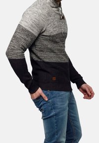 Man som bär en grå och svart stickad tröja med knoppar och blå jeans, med handen i fickan, stående mot en neutral bakgrund.