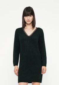 Frau mit glattem, dunklem Haar, die ein schwarzes langärmliges Kleid mit einem Spitzenbesatz am V-Ausschnitt trägt, steht vor einem weißen Hintergrund.