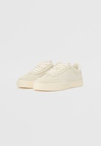 Sneaker beige in suede con una texture liscia, punta rotonda e suola piatta in gomma; logo del brand sul lato; design con lacci; stile minimalista.