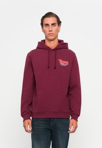 Bordeaux hættetrøje med en frontlomme, der har "Tommy Jeans" logo i kontrasterende farver på brystet. I bomuldsstof, afslappet pasform.