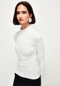 Maglietta bianca a maniche lunghe con tessuto testurizzato, colletto alto e bordi ondulati. La modella indossa grandi orecchini a cerchio e jeans scuri.