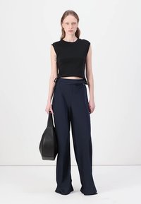 Čierny skrátený top bez rukávov v kombinácii s širokými námorníckymi nohavicami. Modelka drží čiernu tašku. Hladká tkanina, minimalistický dizajn a bočné akcenty.
