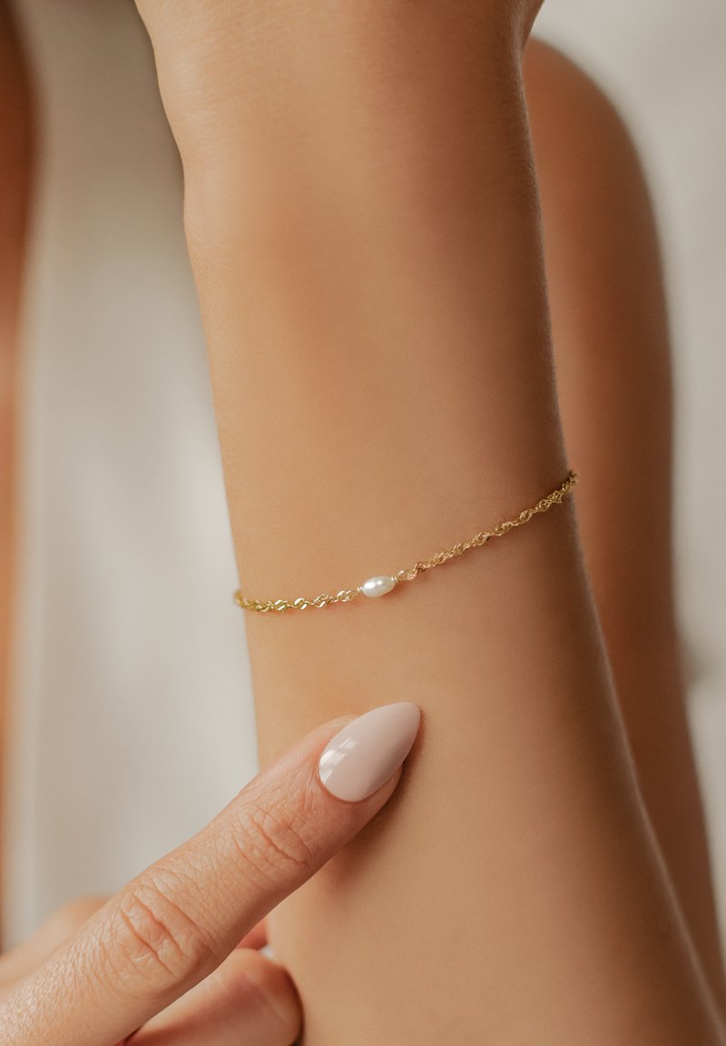 Goldene Kettenarmband mit einem zarten Drehdesign, verziert mit einem kleinen, glatten weißen Perlenakzent, getragen an einem leicht gebräunten Handgelenk.