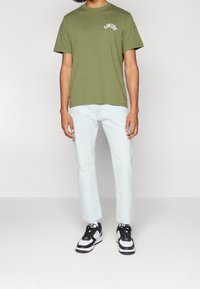 Camiseta de algodón verde oliva con logotipo blanco, combinada con jeans de color azul claro. El calzado incluye zapatillas deportivas negras y blancas.