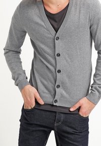 Šedý svetr s véčkovým výstřihem, dlouhými rukávy a šesti tmavými knoflíky. Materiál je hladký a svetr má žebrované manžety a lem.