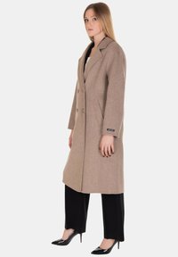 Cappotto beige in lana con design su misura, fronte doppiopetto, rever a lancia, tasche laterali e una texture liscia. Abbinato a pantaloni neri.