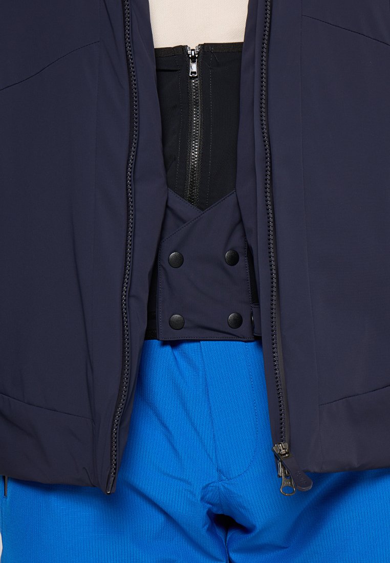 Giacca esterna blu navy con zip sopra una canotta nera stile corsetto, abbinata a pantaloni blu testurizzati con dettagli a pressione e finitura liscia.