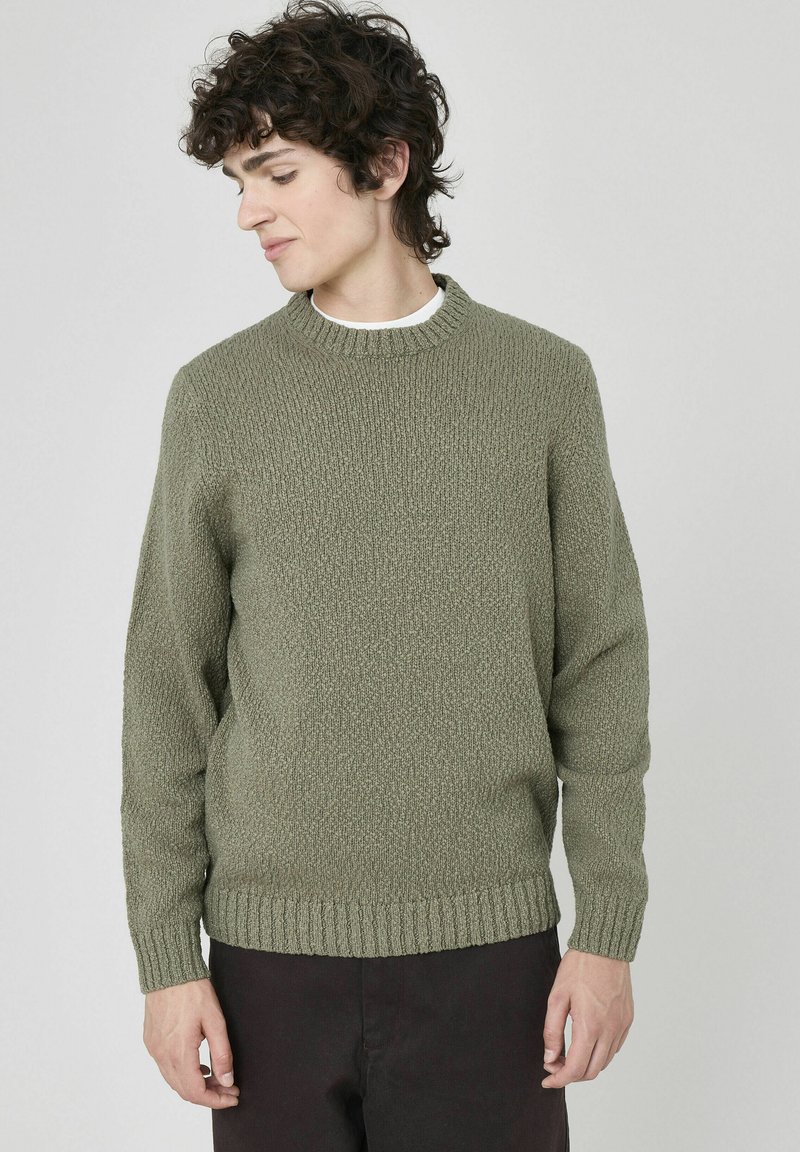 Pull en tricot vert olive avec un col rond et des poignets et un ourlet côtelés. Texture douce et coupe décontractée, associé à un pantalon sombre.