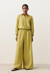 Femeie în picioare, purtând o cămașă galbenă largă, cu nasturi și mâneci lungi, asortată cu pantaloni evazați de aceeași culoare, și șlapi negri.