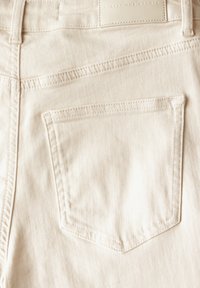 Tela de mezclilla color crema con una sutil textura, que cuenta con un único bolsillo trasero y una pequeña etiqueta grabada en la cintura.