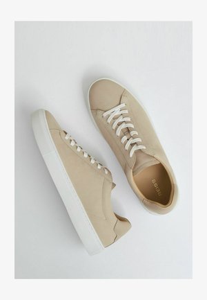 Bežiniai zomšiniai sportbačiai su baltais guminiais padais, turintys plokščias baltas raišteles ir minimalistinį dizainą be papildomų raštų ar akcentų.