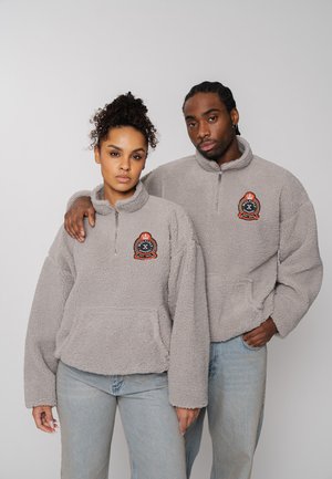 Zwei Erwachsene, die passende graue Fleece-Pullover und hellblaue Jeans tragen, stehen dicht nebeneinander mit ernstem Blick vor einem einfarbigen Hintergrund.