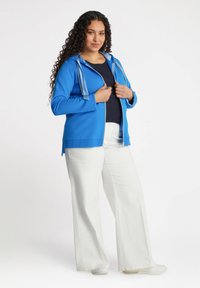 Felpa blu con zip e cappuccio regolabile, maniche lunghe e dettagli bianchi. Abbinata a pantaloni bianchi a gamba larga e calzature chiare.