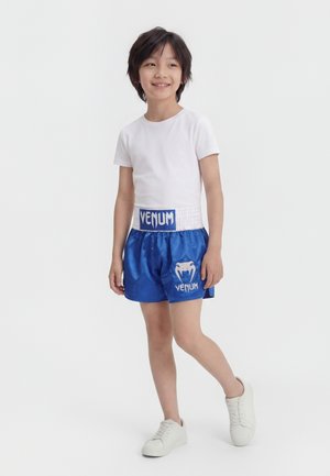 Lächendes Kind mit weißem T-Shirt, blauen Boxshorts mit "Venum"-Logo und weißen Turnschuhen, stehend vor einem schlichten hellen Hintergrund.
