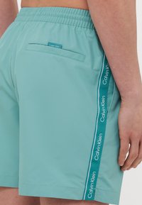 Svetlomodré plavky s textúrovaným povrchom, elastickým pásom, bočnou kapsou a kontrastnými tyrkysovými bočnými pruhmi s brandingom "Calvin Klein".