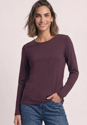 Femme aux cheveux courts et bruns souriante, portant des boucles d'oreilles argentées en forme de créoles, un haut à manches longues violet foncé et un jean bleu avec une main dans la poche.