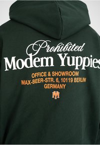 Sweat à capuche vert foncé avec texte blanc et orange indiquant "Prohibited Modern Yuppies" et adresse de bureau à Berlin, Allemagne au dos.
