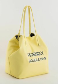 Bolsa de tote amarilla texturizada con dos asas largas, presenta "BOLSA DOBLE AMIGABLE" impreso en texto oscuro en la parte frontal. Forro interior negro.