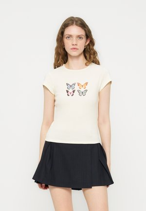 Νέα γυναίκα με κυματιστά μαλλιά που φοράει μια κρεμ μπλούζα με τέσσερις πολύχρωμες πεταλούδες και μια μαύρη πλισέ μίνι φούστα.