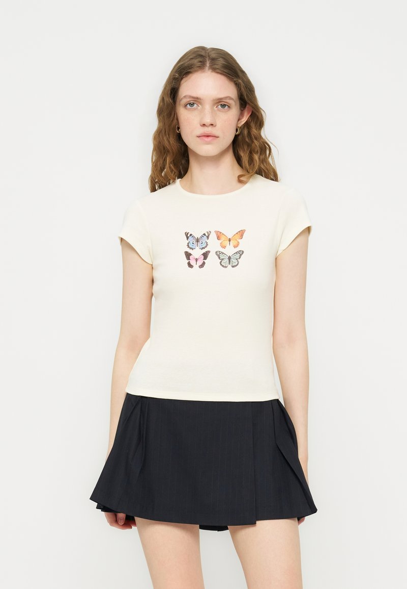 Νέα γυναίκα με κυματιστά μαλλιά που φοράει μια κρεμ μπλούζα με τέσσερις πολύχρωμες πεταλούδες και μια μαύρη πλισέ μίνι φούστα.