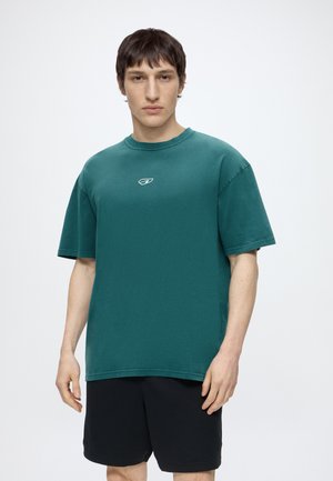 Mladý muž s krátkymi tmavými vlasmi, oblečený v zelenej oversized tričku s malým logom a čiernych šortkách, stojaci na bielom pozadí.