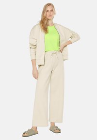 Lightweight beige zip-up jacket, vibrant green t-shirt, loose cream pants, and gray slide sandals with decorative hardware. Casual outfit.