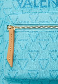 Blå texturerad läderväska med subtil VALENTINO-mönster. Har en guldfärgad dragkedja och ett brun läderdragband på ett framfack.