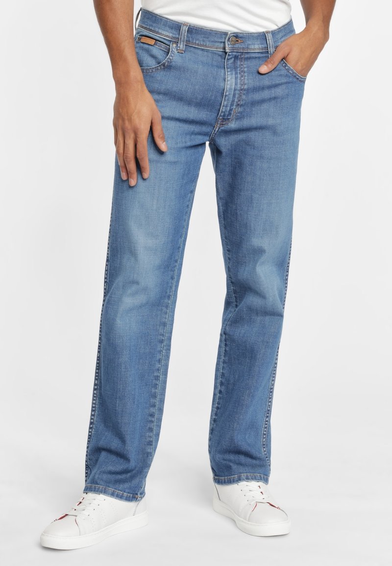 Jeans en denim bleu clair avec une coupe droite, cinq poches, et un patch en cuir marron à la taille ; associés à des baskets blanches.