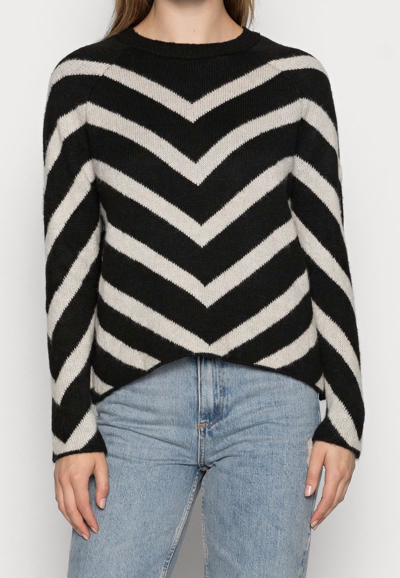 Pull à rayures en chevron noir et blanc, texture tricotée, col rond, manches longues, design court, associé à un jean bleu clair.