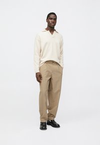 Pull beige en maille avec col en V et col, associé à un pantalon plissé marron clair. Des chaussures noires complètent la tenue.