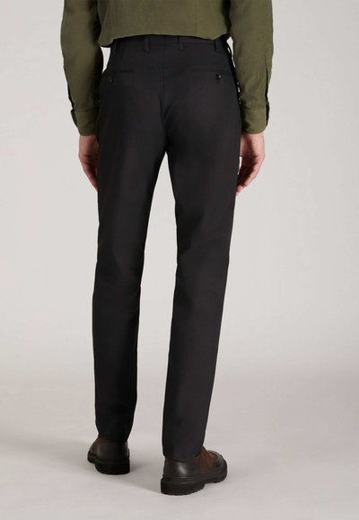 Pantalon noir ajusté, avec une texture lisse et deux poches à l'arrière. Porté avec des chaussures sombres, mettant en avant un design simple.