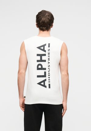 Homme portant un débardeur blanc avec le texte vertical noir "ALPHA INDUSTRIES" au dos, associé à un pantalon noir, de dos.