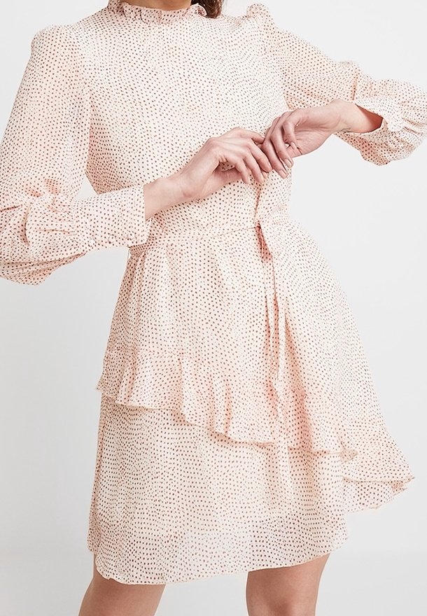 Femme portant une robe rose clair à manches longues avec un petit motif à pois et une jupe à volants superposés, mains jointes à la taille.