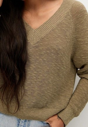 Person iført en tekstureret beige V-hals sweater med langt mørkt hår over skulderen og hånden stukket ned i lommen på lyseblå jeans.