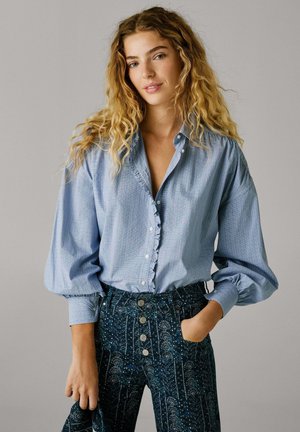 Svetlo modra bluzica s gumbi in nabranim robom, v kombinaciji z visokimi, vzorčastimi temnimi hlačami, ki imajo kovinske gumbe in cvetlične vzorce.